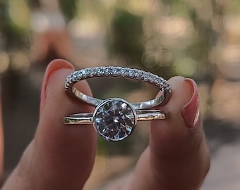 May include: Two rings, a set, are held up against a blurred background. The first ring has a large, round, clear stone set in a bezel. The second ring is a band with small, clear stones. Both rings have a gold band.