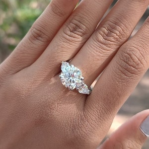 Puede incluir: Primer plano de una mano con un anillo de plata con una piedra central grande, redonda y transparente y dos piedras más pequeñas en forma de pera a cada lado.