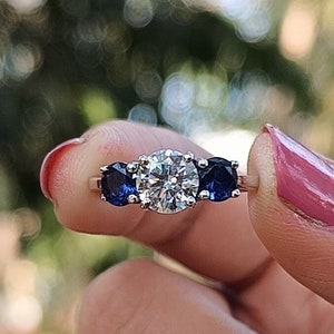 May include: A gold ring with a large, clear, round central stone and two smaller, dark blue stones. The ring is held up against a blurred green background.