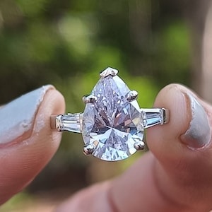 May include: A pear-shaped engagement ring with a large, clear, cubic zirconia stone set in a silver band. The ring has two smaller cubic zirconia stones on either side of the main stone.
