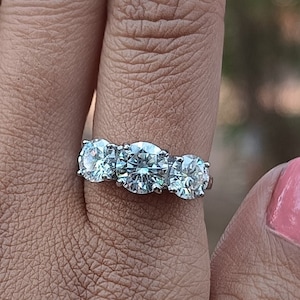 May include: A silver ring with three large round diamonds set in a band. The ring is on a person's finger.