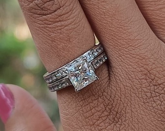 Puede incluir: Un anillo de plata con una piedra preciosa grande, de corte cuadrado y transparente. El anillo tiene una banda de piedras preciosas más pequeñas y transparentes.