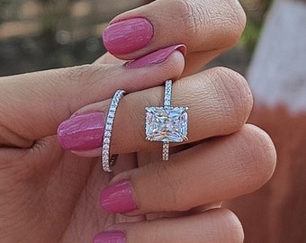 Puede incluir: Primer plano de una mano con un anillo de plata con un gran diamante de forma cuadrada. El anillo está engastado con diamantes más pequeños alrededor de la piedra central. La mano también lleva una fina banda de plata en el mismo dedo.