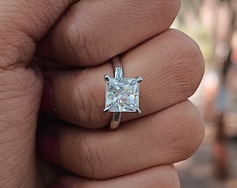 May include: A silver ring with a large, square-shaped, clear gemstone. The ring is on a person's finger.