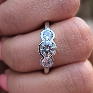 Puede incluir: Un anillo de plata con tres diamantes redondos engastados en fila. El anillo está en el dedo de una persona.