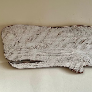 May include: White painted wooden whale wall decor. The whale is in a simple silhouette shape with a slightly curved body and a small tail fin.