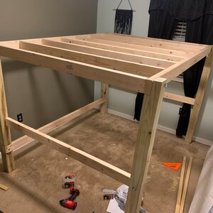 以下が含まれることがあります： ベージュの壁とカーペットのある部屋で、ロフトベッドのウッドフレームが組み立てられています。フレームは未仕上げのウッド製で、4本の柱で支えられています。フレームは部分的に組み立てられており、上部と下部のレールが設置されています。フレームの周りには工具やその他の材料が散らばっています。
