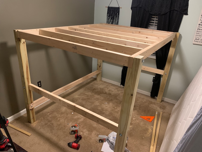 以下が含まれることがあります： ベージュの壁とカーペットのある部屋で、ロフトベッドのウッドフレームが組み立てられています。フレームは未仕上げのウッド製で、4本の柱で支えられています。フレームは部分的に組み立てられており、上部と下部のレールが設置されています。フレームの周りには工具やその他の材料が散らばっています。