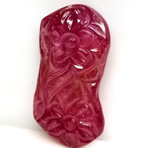 May include: A carved, polished gemstone in shades of pink and red. The stone features a floral design with intricate details. The shape is irregular, with a smooth, glossy surface. The gemstone is set against a white background.
