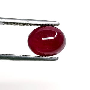 Könnte beinhalten: Ein oval geschliffener, tiefroter Edelstein, der von einer silbernen Pinzette gehalten wird. Der Stein hat eine glatte, glänzende Oberfläche und eine satte Farbe. Der Hintergrund ist schlicht weiß, was den Farbton des Steins hervorhebt.