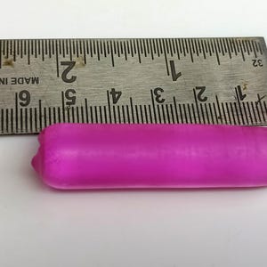 Può includere: Un oggetto cilindrico rosa acceso è posizionato accanto a un righello di metallo. Il righello, segnato in centimetri, indica la lunghezza dell'oggetto. L'oggetto ha una consistenza liscia e sembra essere fatto di un materiale morbido.