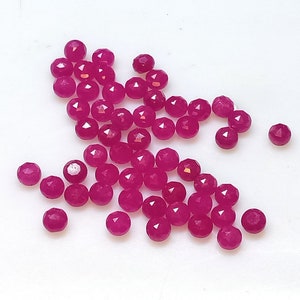 May include: A collection of faceted, round, magenta-colored gemstones scattered on a white surface. The gemstones have multiple facets that catch the light, creating a sparkling effect. These small, polished stones are likely used for jewelry making.