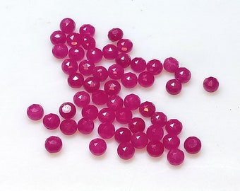May include: A collection of faceted, round, magenta-colored gemstones scattered on a white surface. The gemstones have multiple facets that catch the light, creating a sparkling effect. These small, polished stones are likely used for jewelry making.