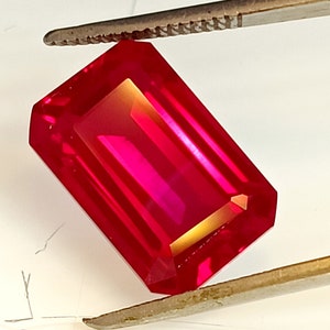 Op de afbeelding: Een levendige rode edelsteen met een rechthoekige smaragd-slijpvorm. De edelsteen wordt vastgehouden door een pincet.