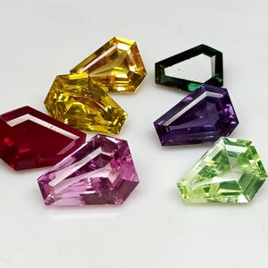 May include: Seven multi-coloured, coffin-cut gemstones. The gems are in shades of yellow, green, purple, pink, and red. Each stone has a flat top and angled sides. The gemstones are displayed on a white surface.
