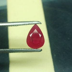 May include: A teardrop-shaped, deep red gemstone held by tweezers.