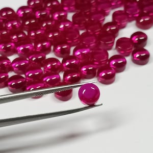 May include: A close-up of a pile of bright pink round cabochon gemstones. One gemstone is being held by tweezers.