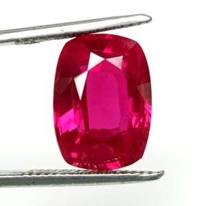 May include: A vibrant, deep red gemstone with a cushion cut. The gemstone is held in a pair of tweezers.