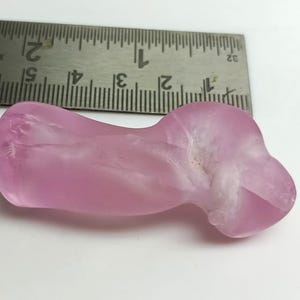 May include: A translucent, light pink gemstone with an irregular, organic shape. A metal ruler with inch markings is placed above the stone. The stone appears to be approximately 2 inches in length.