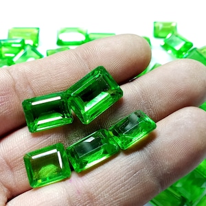 Könnte beinhalten: Eine Sammlung von smaragdgrünen Edelsteinen, die in rechteckige Formen geschnitten sind, wird auf einer Hand präsentiert. Die Edelsteine sind facettiert und haben eine leuchtende, lebendige Farbe.