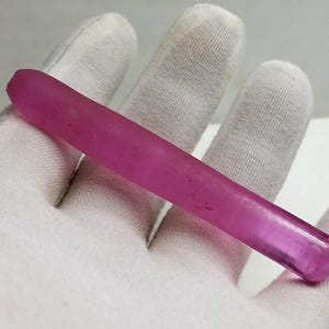 Peut inclure: Une pierre précieuse rose, allongée et polie, tenue dans une main gantée. La pierre a une forme cylindrique avec une extrémité arrondie et une pointe facettée. La couleur de la gemme est rose foncé et translucide.