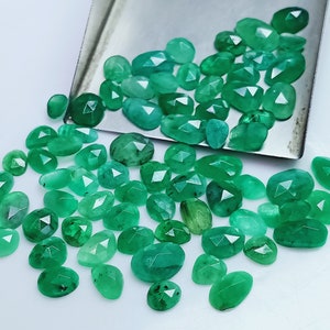 Könnte beinhalten: Eine Sammlung grüner Smaragdsteine, in verschiedenen Formen und Größen geschnitten, auf einer weißen Oberfläche verstreut. Die Edelsteine sind facettiert und haben eine polierte Oberfläche.