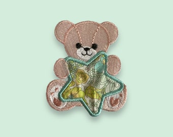 May include: A pink embroidered teddy bear holding a green star-shaped fabric with a floral pattern.