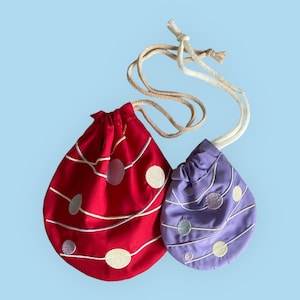 Puede incluir: Dos bolsas de cordón con asas de cuerda blanca. La bolsa más grande es roja con círculos y líneas blancas bordadas. La bolsa más pequeña es morada con círculos y líneas blancas bordadas.