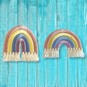 以下が含まれることがあります： パステルカラーの刺繍された虹のパッチが2つ。各パッチは、ピンク、黄色、青、紫のアーチと、下部に白いフリンジが特徴です。青い木製の背景に配置されています。