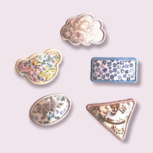 Op de afbeelding: Vijf stoffen patches met verschillende ontwerpen. De patches zijn in de vorm van een wolk, een rechthoek, een ovaal, een driehoek en een berenkop. De patches zijn allemaal versierd met bloemmotieven in tinten roze, blauw en geel.