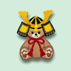 Könnte beinhalten: Ein brauner, gestickter Teddybär, der einen gelben und schwarzen Samurai-Helm mit einer roten Schleife trägt.