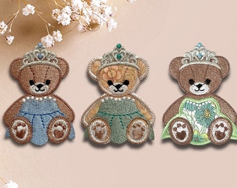 May include: Three embroidered patches of brown teddy bears wearing princess crowns and dresses. The dresses are blue, green, and floral.