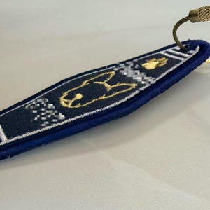 May include: A dark blue fabric keychain tag featuring gold embroidered outlines of a dog head and a paw print. The tag has white embroidered stripes at the top and bottom, with a metallic gold screw-lock cable ring attached to the top corner.