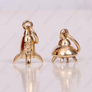 Pode incluir: Dois pingentes dourados, um com a forma de um foguete e o outro com a forma de um OVNI, são mostrados em um fundo branco. Ambos os pingentes têm um pequeno laço na parte superior para serem presos a um colar ou pulseira.