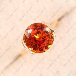May include: A round, orange gemstone set in a gold ring. The gemstone is faceted and has a bright, fiery orange color.