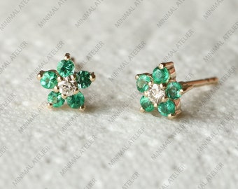 May include: A pair of gold earrings with a flower design. Each earring features a small diamond in the center surrounded by six emerald green stones.