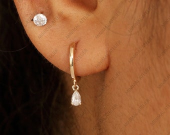 May include: A pair of gold hoop earrings with a teardrop-shaped diamond dangling from one earring. The earrings are shown on a person's ear.