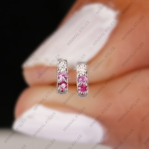 May include: A pair of small hoop earrings with a row of pink and white gemstones. The earrings are made of silver and are a delicate and minimalist design.