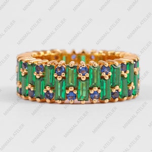 Könnte beinhalten: Ein Goldring mit smaragdgrünen und blauen Baguette-Edelsteinen. Der Ring ist in eine Goldschiene gefasst und hat eine polierte Oberfläche.