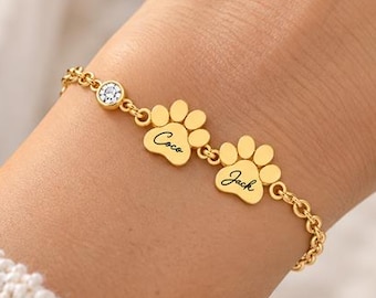 May include: A gold-toned chain bracelet featuring two paw print charms engraved with the names Coco and Jack, alongside a single small round clear stone accent. The bracelet is secured with a delicate link chain design.