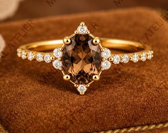 May include: A gold ring featuring a central oval-cut smoky brown gemstone surrounded by a halo of small round white stones. The band is thin and metallic, with additional small white stones set along the sides of the central setting.