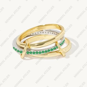 Può includere: Un anello dorato con più fasce, alcune decorate con piccole gemme verdi scintillanti. L'anello ha un design moderno e intrecciato, con il testo "MINIMAL ATELIER" ripetuto sullo sfondo.