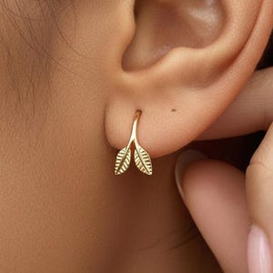 May include: A gold hoop earring featuring a small, delicate branch design with two textured leaves. The earring is shaped like a curved stem that splits into two pointed leaf shapes, each with fine etched lines detailing the leaf veins.