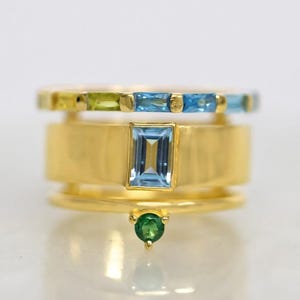 May include: A gold ring with a stack of three bands. The top band has a row of light blue and green gemstones. The middle band has a larger light blue gemstone. The bottom band has a small green gemstone.
