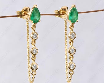 May include: A pair of gold earrings featuring a pear-shaped green gemstone stud connected to a dangling chain with three small round clear stones. The earrings are suspended from a thin wire, and the text MINIMAL ATELIER is visible.