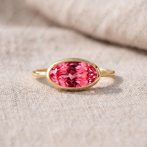 May include: A gold ring featuring a horizontal oval-cut pink gemstone set in a smooth bezel setting. The band is thin and polished, showcasing a vibrant, faceted pink stone that reflects light across its surface. The design is simple and elegant.