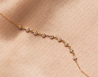 May include: A delicate gold necklace with a series of small, round, clear gemstones. The gemstones are evenly spaced along the necklace's chain. The necklace is displayed on a soft, peach-colored fabric.