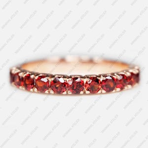 May include: A delicate gold band ring with a row of small, round, red gemstones. The ring is a classic and elegant design.