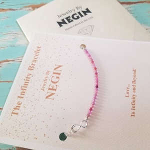 Könnte beinhalten: Ein pinkfarbenes Edelstein-Unendlichkeitsarmband mit einem silbernen Unendlichkeitsanhänger. Das Armband befindet sich auf einer weißen Karte mit dem Text "The Infinity Bracelet Jewelry By NEGIN Love... To Infinity and Beyond!"