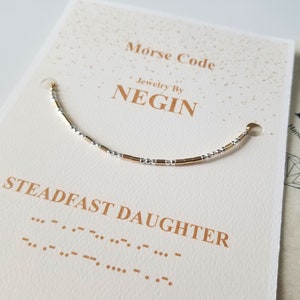 Op de afbeelding: Een armband met gouden en zilveren kralen met de woorden "Steadfast Daughter" en de bijbehorende morsecode eronder. De armband ligt op een witte kaart met de woorden "Morse Code Jewelry by Negin" erop gedrukt.